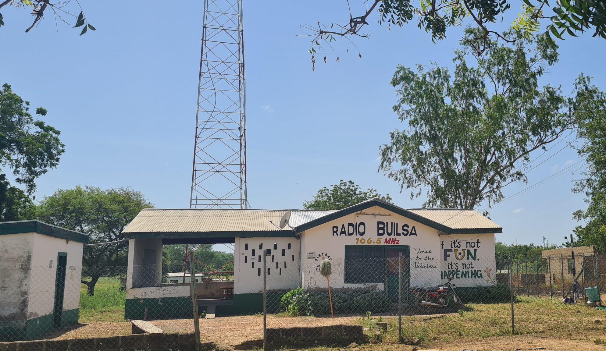 Hausfront von Radio Builsa mit dem Schriftzug „Radio Builsa“ in Schwarz und „106.5 MHz“ in Gelb und Rot über der Tür. Rechts steht der Spruch „If it's not FUN it's not HAPPENING“ in fröhlichen Farben (Schwarz, Gelb, Grün). Im Hintergrund ist der Sendemast teilweise sichtbar. Das Bild erscheint im News-Beitrag Radio Builsa's Solar Repairs